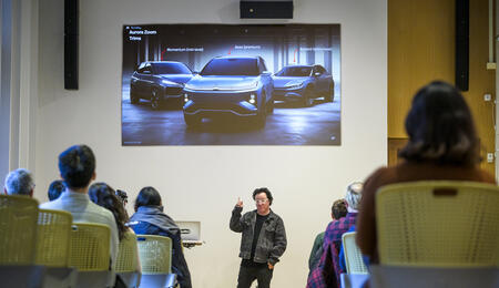 A person giving a presentation to a room full of people. A photo of three cars is projected on a screen above him.