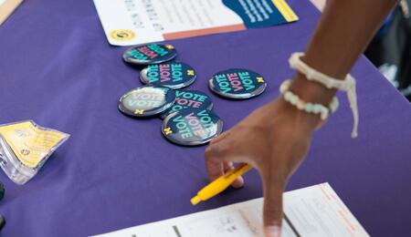 A pile of UMichVotes buttons sit on a table near someone filling out a form.