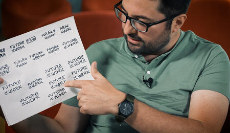 Pedro Pazmino holds up a sheet of paper covered in hand drawn early versions of his Future of Work logo. He points to one.