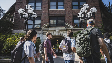 Students wearing backpacks walk on a sidewalk through U-M's Ann Arbor campus. One backpack has a UMSI logo