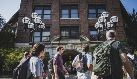 Students wearing backpacks walk on a sidewalk through U-M's Ann Arbor campus. One backpack has a UMSI logo