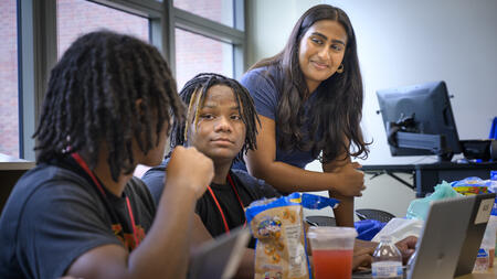A teaching assistant stands leans over a desk engaging with two students 