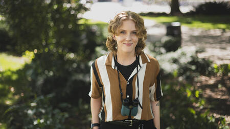 Meg Millure stands in a green area on campus with their hands in their pockets, wearing binoculars around their neck