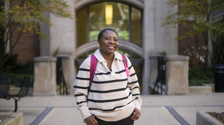 Olga Hamilton smiles in front of the entrance to a campus building, wearing a pink backpack