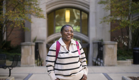 Olga Hamilton smiles in front of the entrance to a campus building, wearing a pink backpack