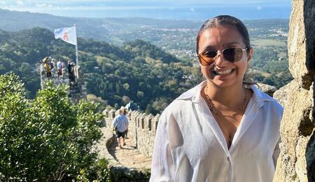 Jessica Thomas smiling standing on stone wall with turret and flag at its end. Background of rolling tree-covered mountains.