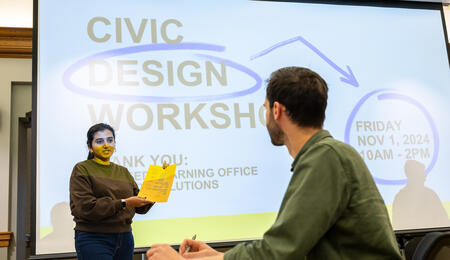 A student holds a piece of paper in front of a screen that reads Civic Design Workshop, while another student looks on