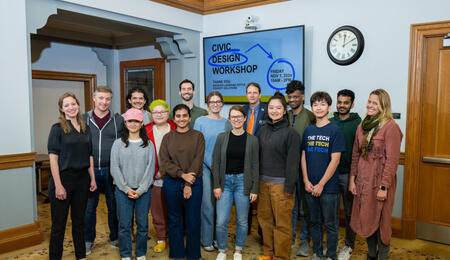 A group of sixteen participants and organizers from the civic workshop pose in front of a lobby monitor that says "Civic Design Workshop"
