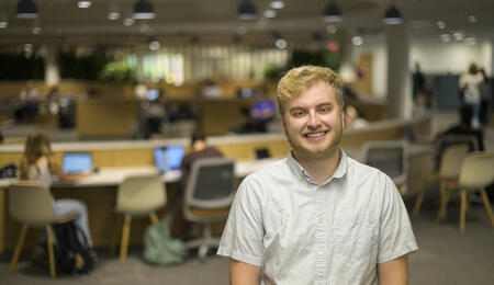 Austin Book poses in the library with out-of-focus students studying behind him