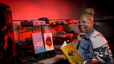 Kishonna sitting in front of a video game, mid-laugh and holding a copy of her book, Woke Gaming. She is surrounded by red light. 