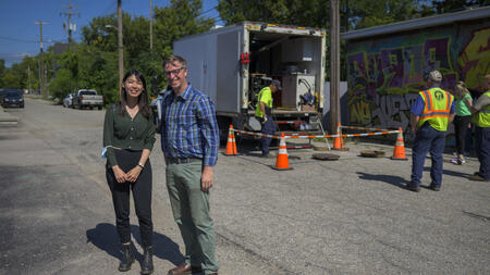 A student and lecturer stand in an alleyway with a white truck, a few public service professionals, and orange cones visible behind them