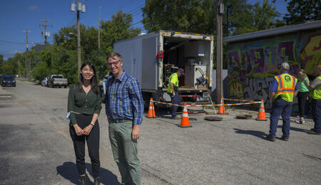 A student and lecturer stand in an alleyway with a white truck, a few public service professionals, and orange cones visible behind them