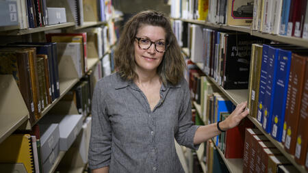 A librarian stands in the aisle between library shelves, with one arm resting on a book