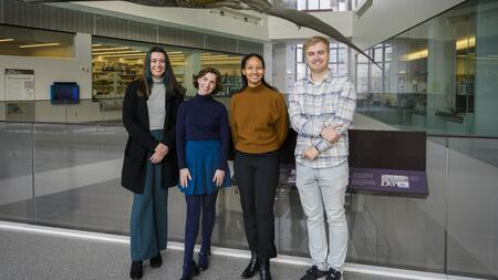 Madison Haack, Julia Cave Arbanas, Cameron Robertson and Austin Book pose on the second level of a museum with a life-size model of a prehistoric pterosaur visible behind them