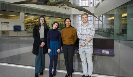 Madison Haack, Julia Cave Arbanas, Cameron Robertson and Austin Book pose on the second level of a museum with a life-size model of a prehistoric pterosaur visible behind them