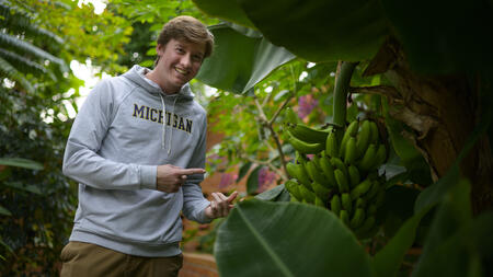 A student wearing a University of Michigan hoodie points at a plant in a greenhouse.