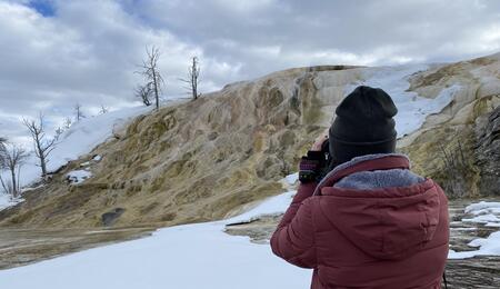 An individual in a red jacket, seen from behind, looks through binoculars at the snow-covered Mammoth Hot Springs