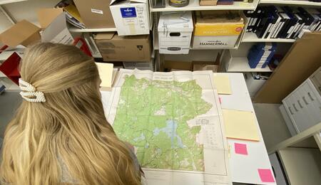 The back of a student's head as she studies a large map laid out on a table in front of her, with manuscript boxes visible on shelves in front of her