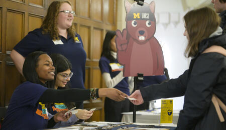 UMSI staff members sitting behind a table and handing stickers to students. Next to the table is a large cutout of a squirrel wearing a UMSI beanie.