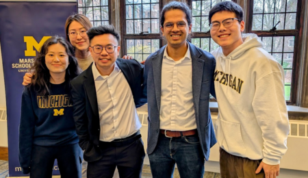 Five students pose, smiling, in a bright classroom setting with a banner behind them that displays the Michigan block M