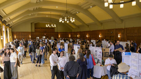 A view of a crowd gathered in a well-lit ballroom with tall ceilings, filled with tables holding posters 
