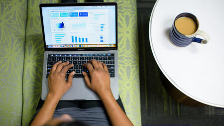 A view from above as a person works on a laptop with graphs visible on the screen. A coffee mug rests on a white table beside them