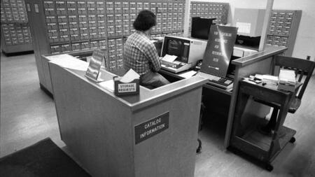 A black and white photo of an individual sitting at a reference desk with an old computer. A wall is lined with card catalogs, and a sign on the desk reads "Catalog information." Another sign with an arrow reads "For catalog assistance ask at reference desk" 