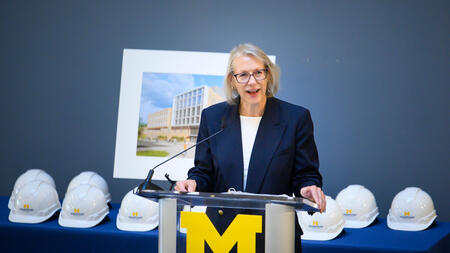 Yakel speaks at a podium adorned with a block M. Behind her, a table is filled with white construction hats and a framed rendering of a building