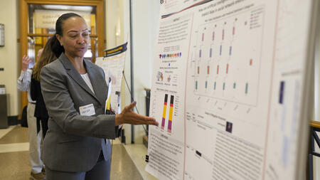 A person wearing a gray suit gestures at a large display poster with small text and data visualizations