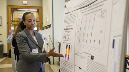 A person wearing a gray suit gestures at a large display poster with small text and data visualizations
