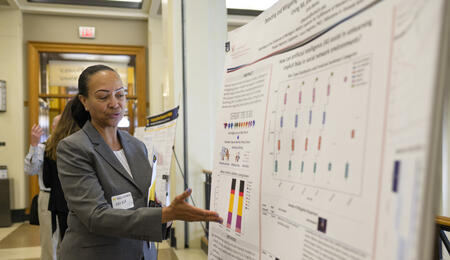A person wearing a gray suit gestures at a large display poster with small text and data visualizations