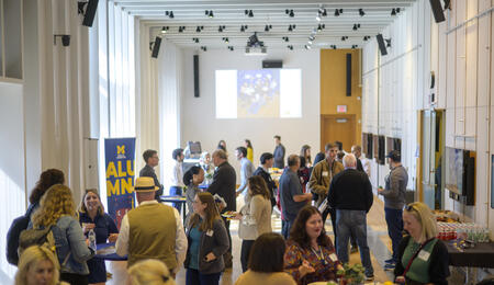Many people gather in a bright event space with round tables. A banner reads "Alumni," and a slideshow of photos is projected on the back wall