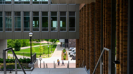 View through an architectural overpass connecting two buildings, looking down onto a pathway with three people walking and verdant campus grounds in the background.