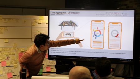 Ki Hyun Ahn gestures at a prototype displayed on a screen, with students' heads visible in the foreground and a whiteboard covered in PostIt notes behind him