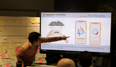 Ki Hyun Ahn gestures at a prototype displayed on a screen, with students' heads visible in the foreground and a whiteboard covered in PostIt notes behind him