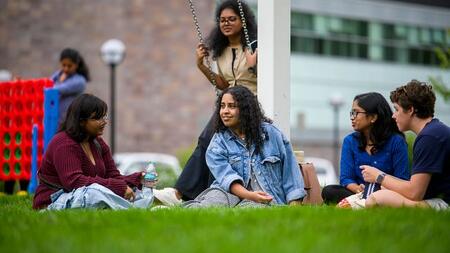 Four people sit on the grass and chat. A person behind them is sitting on a swing.