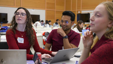 Three teammates, all wearing red, smile and glance to the left. Each has a laptop on a table in front of them in a large academic room
