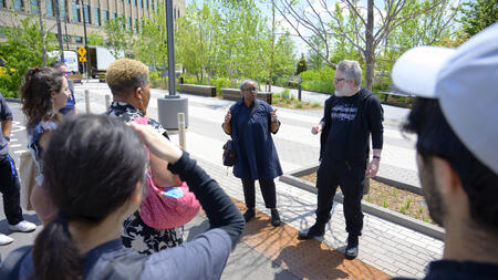Cezanne Charles and John Marshall stand on a sidewalk in front of greenery, addressing a small tour group