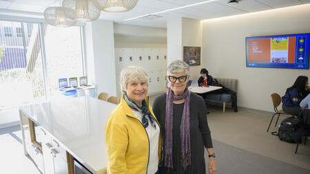 Sue Quackenbush and Kenlee Ray posing in the student lounge, with a wide kitchen island and hanging light fixtures behind them. Students seated at tables and a row of lockers are visible in the background.