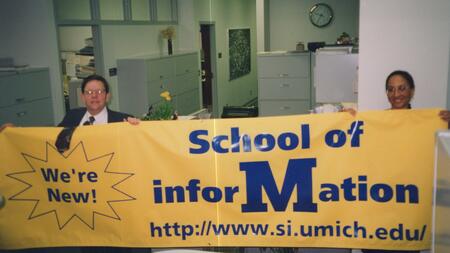 Daniel E. Atkins and C. Olivia Frost hold up a large maize promotional banner that says "We're new! School of Information."