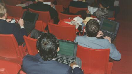 The backs of several students' heads as they work on old Apple laptop hops in an orange velvet lecture hall