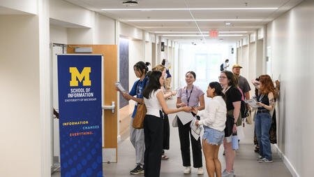 A group of students gather, facing each other and chatting, in a brightly lit hallway with a tall blue UMSI pull-up banner that says Information changes everything