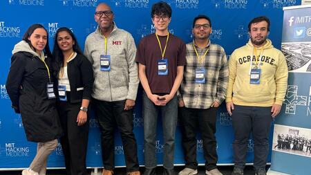 Simranjeet Singh poses with fellow hackathon participants in front of a backdrop with repeated MIT Hacking Medicine logos. Everyone wears nametag lanyards.
