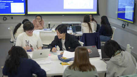 Students work together at a table in a brightly lit classroom. One smiles while leaning over to a student to their left