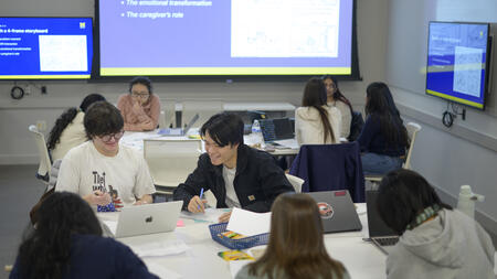 Students work together at a table in a brightly lit classroom. One smiles while leaning over to a student to their left