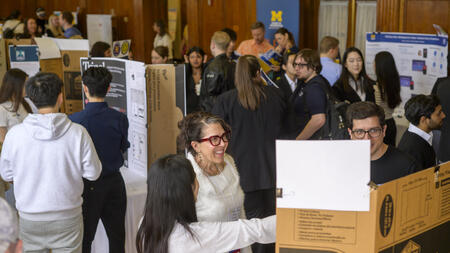 Many people gather in a large room around different posters, with a person gesturing to a poster in the foreground