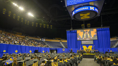 Rows of graduates face the stage on the brightly lit floor of the Crisler Center