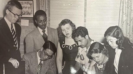 Black-and-white archival photo of seven people gathered around a table, reviewing papers and documents together. One person writes while others stand or sit closely around the materials.