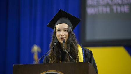 Alyssa Peck speaks into a microphone wearing a graduation cap and gown, with part of a screen visible behind her