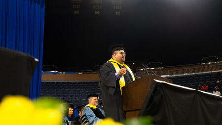 Patrick Mullen-Coyoy stands at a podium, with yellow flowers visible in the foreground lower than the stage